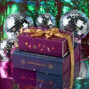 Stacked Woops! New Year’s macaron gift boxes with gold ribbons surrounded by shiny disco balls and a colorful reflective backdrop.