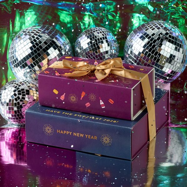 Stacked Woops! New Year’s macaron gift boxes with gold ribbons surrounded by shiny disco balls and a colorful reflective backdrop.