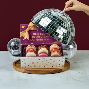 Woops! New Year’s macaron gift box with colorful macarons on a wooden tray, partly covered by a disco ball lid, with more disco balls and festive decor in the background.