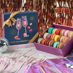 Woops! New Year’s macaron gift box with colorful macarons beside festive packaging featuring champagne glasses, surrounded by disco balls and metallic decorations.