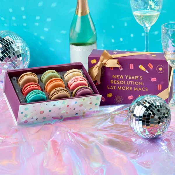 Woops! New Year’s macaron gift boxes with gold ribbons displayed on a shiny surface surrounded by disco balls, champagne glasses, and festive decorations.