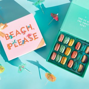 A light blue background features a Woops! box full of 18 assorted French macarons and a macaron box with a pink Summer sleeve to its left. Some cocktail umbrellas are lying around.