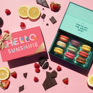 A light pink background features a box of 9 French macarons with assorted flavors and a macaron box with a summer sleeve to the left. Some fruits and chocolates are lying around.