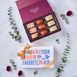 A box with nine French macarons has a macaron box with a Mother's Day sleeve below. Surrounding them are branches with green leaves.