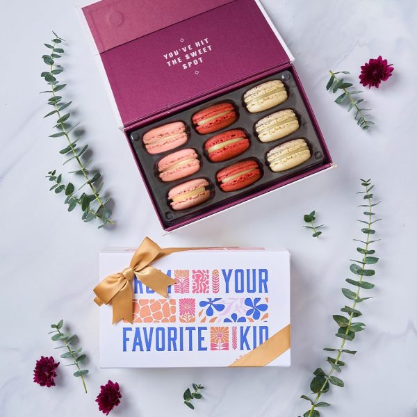A box with nine French macarons has a macaron box with a Mother's Day sleeve below. Surrounding them are branches with green leaves.