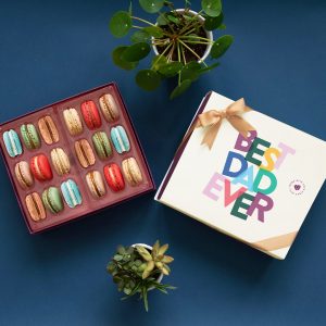 A box full of 18 assorted French macarons has a French macaron box with a Father's Day sleeve to its right. Above and below them are some plants.