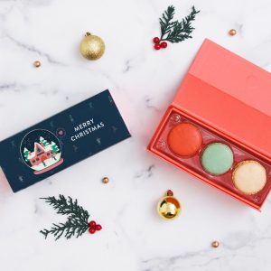 A box full of three French macarons has a macaron box with a blue Holiday sleeve to the left. Lying around are numerous holiday ornaments.