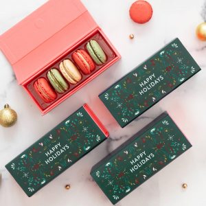 A box full of three French macarons has a macaron box with a blue Holiday sleeve to the left. Lying around are numerous holiday ornaments.