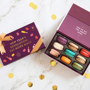 A box full of nine assorted macarons has a macaron box with a purple New Year's sleeve to the left. Surrounding them is golden confetti.