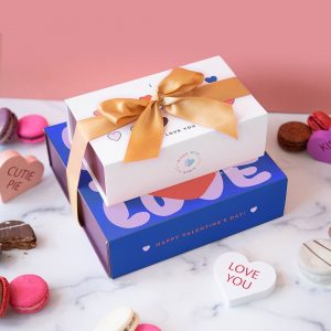 A stack of two macaron boxes with Valentine's Day sleeves is surrounded by assorted macarons.