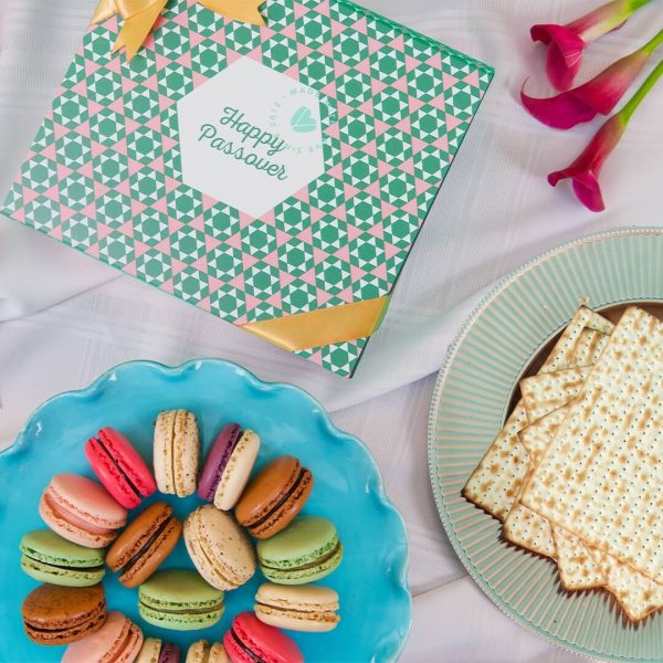 A macaron box with a colorful Passover sleeve has a plate full of assorted macarons and a plate full of matzos below.