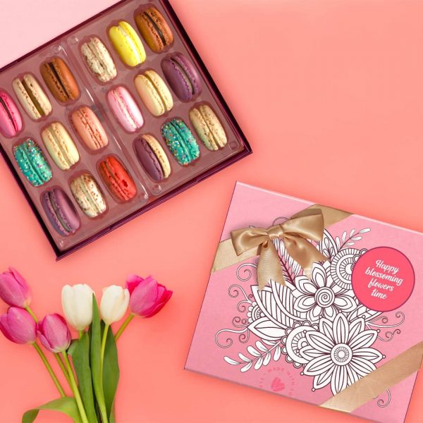 A box full of 18 assorted French macarons has a macaron box with a Blossoming Colors sleeve and some tulips below.