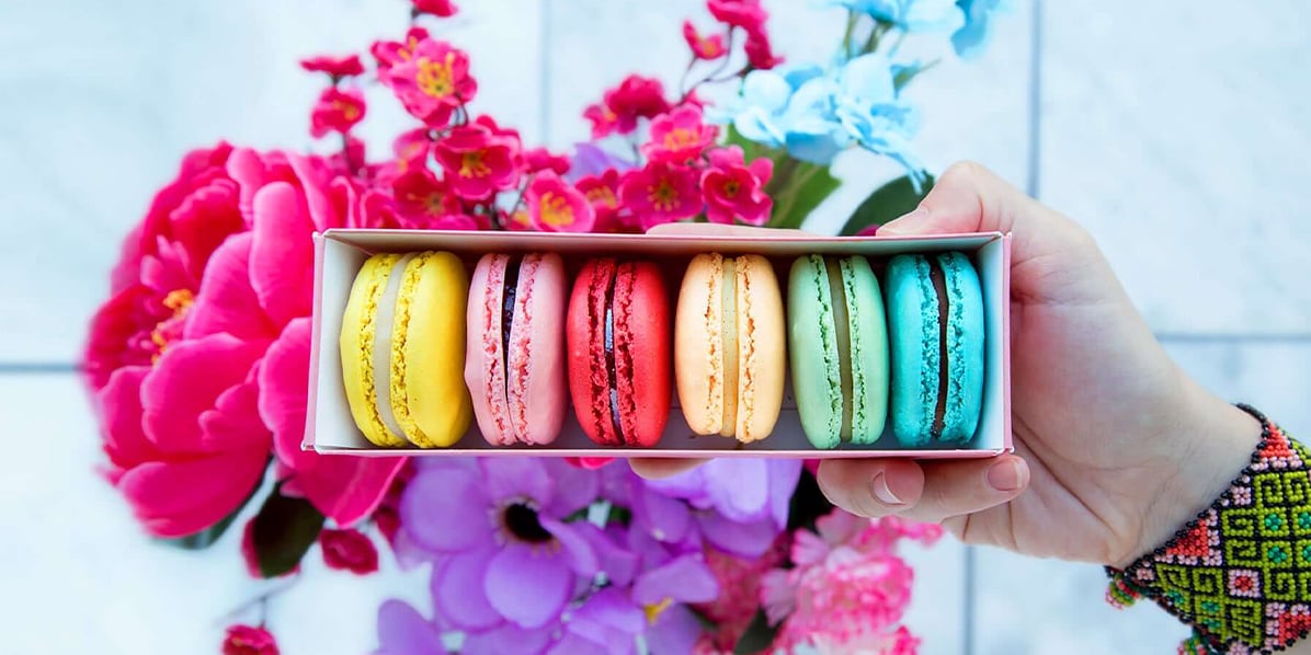 Box of 6 macarons being hold by a woman’s hand with colorful bracelet and flowers in the background