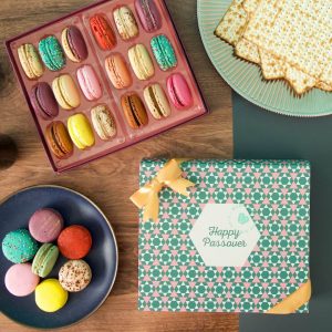 A macaron box with a colorful Passover sleeve has a box full of assorted macarons above. To the left is a plate full of assorted macarons and to the right a plate with matzos.