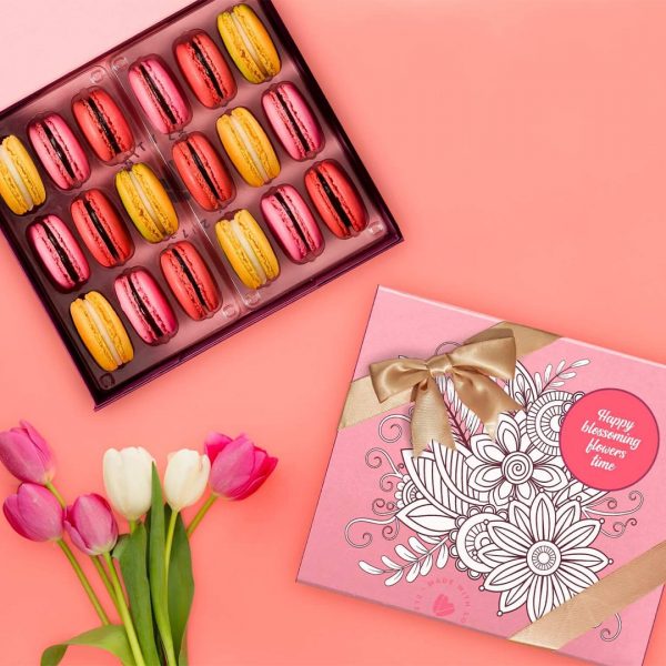 A box of 18 French macarons with Strawberry Lemonade, Raspberry, and Lemon Meringue macarons has a box of French macarons with a pink and white Blossoming Colors sleeve to its side. Below are some pink and white tulips.