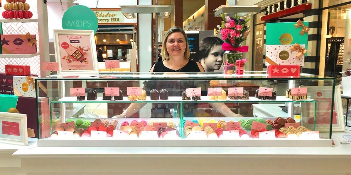 Blonde woman smiling behind a Woops! boutique kiosk filled with French macarons