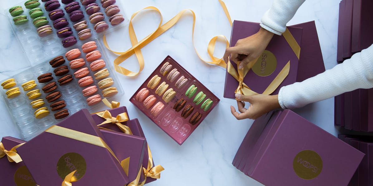 Several Woops! purple French macaron boxes and some assorted macaron trays with hands tying a ribbon