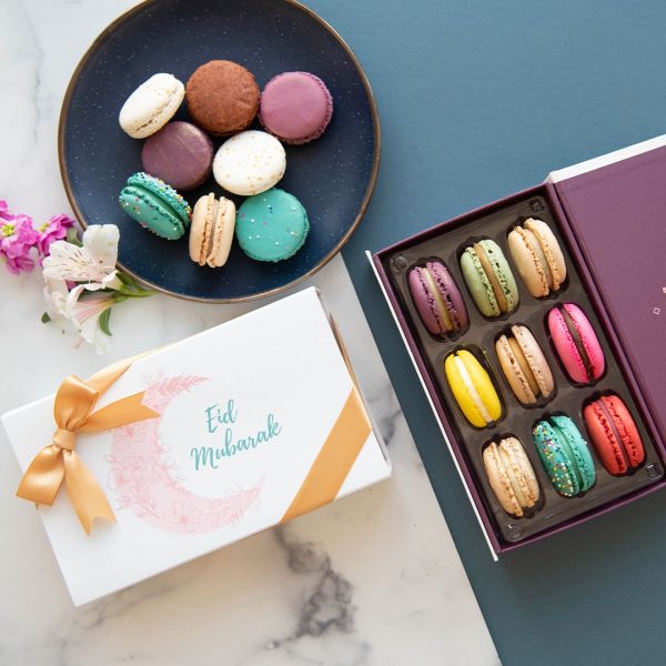 A box full of nine assorted macarons has a macaron box with a light blue Eid sleeve to the left. At the top is a plate full of assorted French macarons.