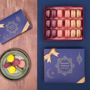 A box full of 18 assorted macarons has two macaron boxes with dark blue Ramadan sleeve to the left and below. On the bottom left corner is a plate full of assorted macarons.