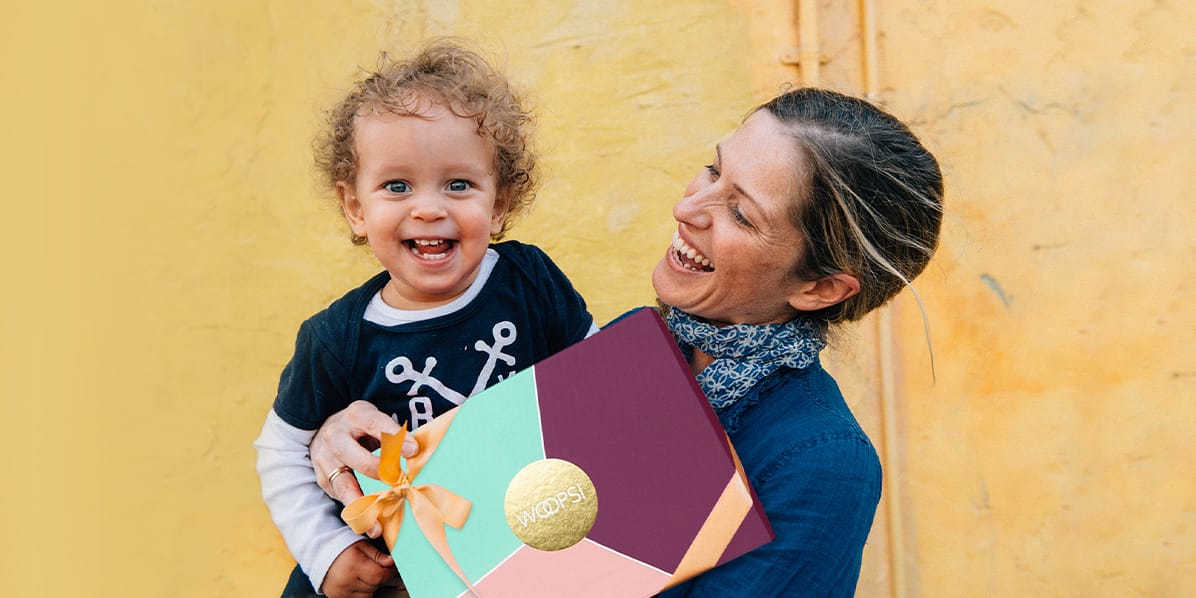 A yellow background features a smiling mother with a dark blue shirt and blue scarf looking at her baby that she’s holding in her arms along with a pink with golden ribbon Woops! macarons box. The baby has a black & whited shirt, curly light hair and is smiling, as well.