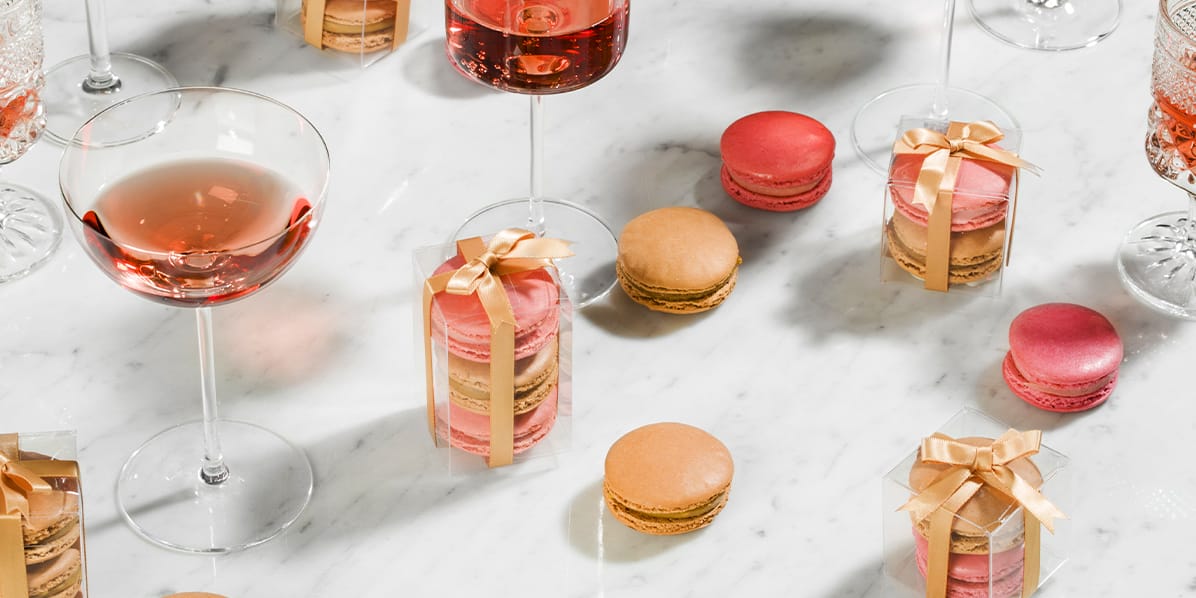 Cups of pink champagne are surrounded by macaron favor boxes with golden ribbons and single macarons on a marble countertop