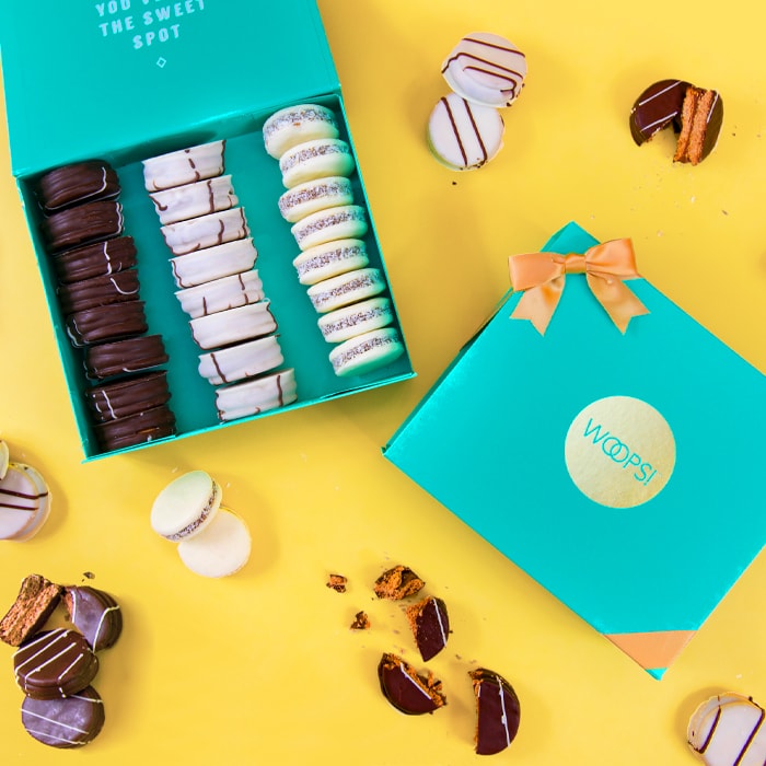 A yellow background features an open Woops! French macarons box full of assorted alfajores. To the right is a closed aquamarine Woops! French macarons box with a golden ribbon and several alfajores lying around.
