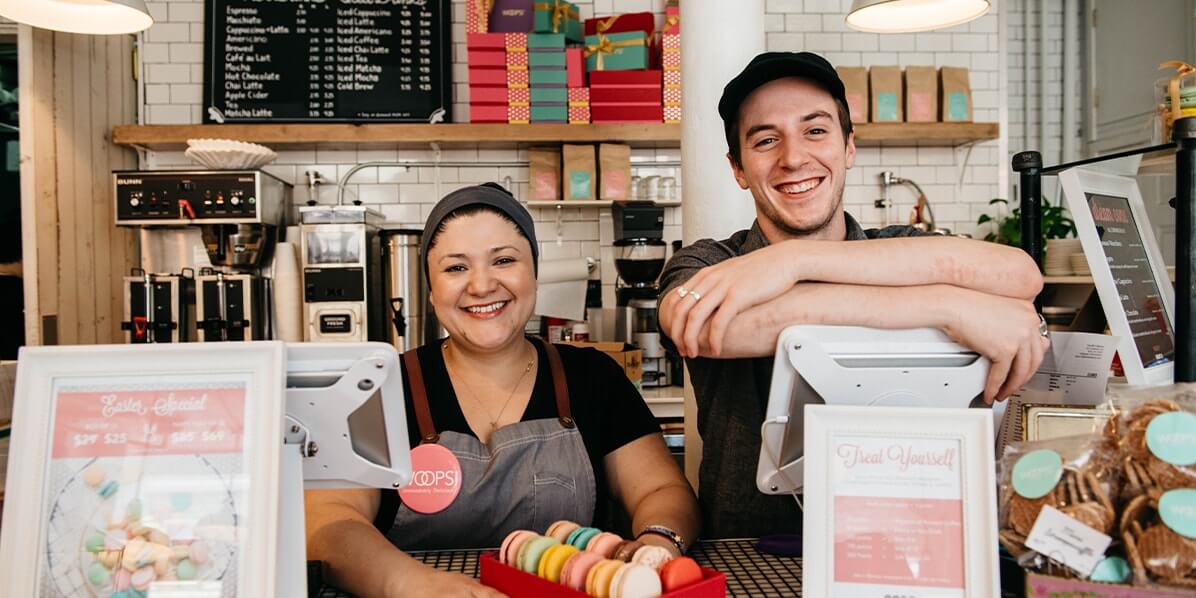 A smiling lady and a smiling guy are looking at the camera behind a Woops! Bakeshop counter. The lady has a red Woops! box of 12 French macarons in assorted flavors.