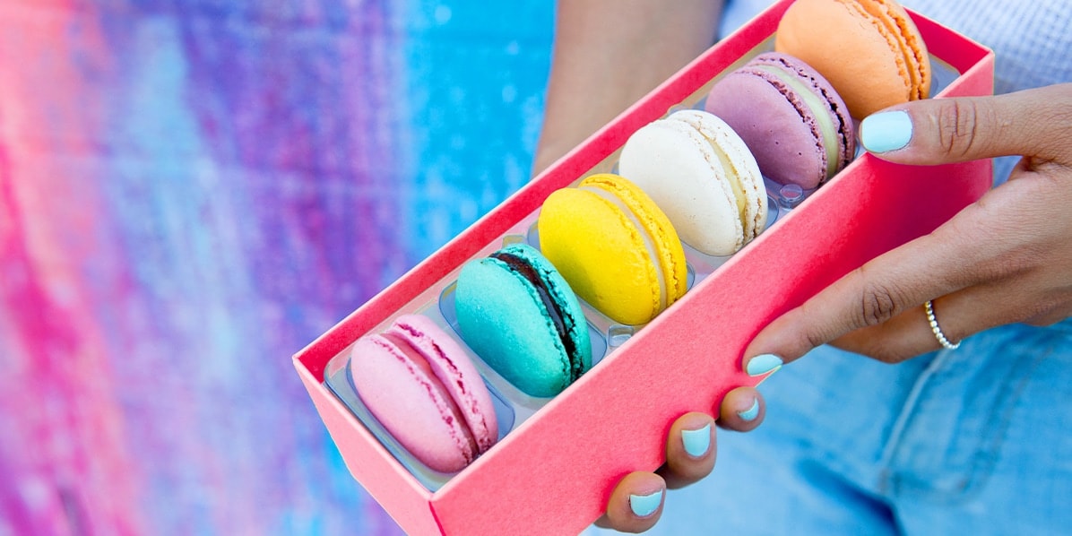 Yellow Woops! Box of 6 French macarons with assorted flavors being held by a female hand and a colorful wall in the background