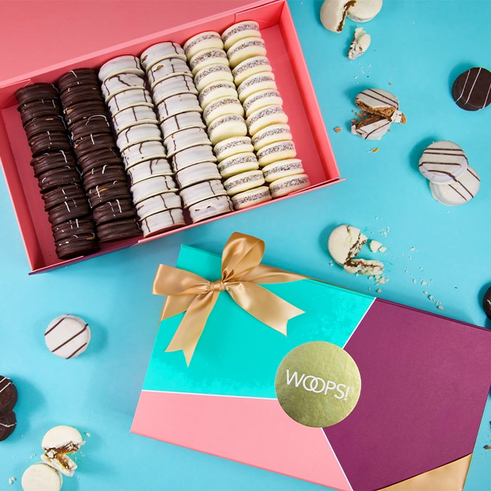 A bright blue background features a Woops! French Macarons box full of alfajores in assorted flavors at the top and below is a closed green, purple, and pink Woops! box with a golden ribbon. Several macarons are lying around.