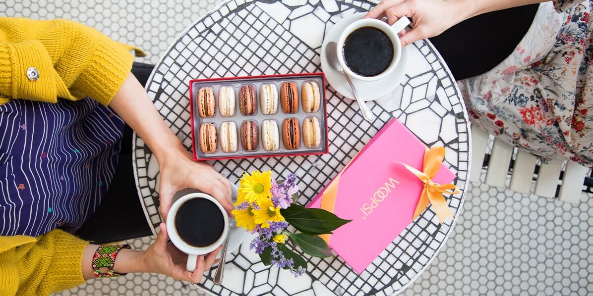 Two women with coffee cups in hand are sharing a Woops! Box of 12 French macarons while sitting at opposite sides of a black & white table with flowers