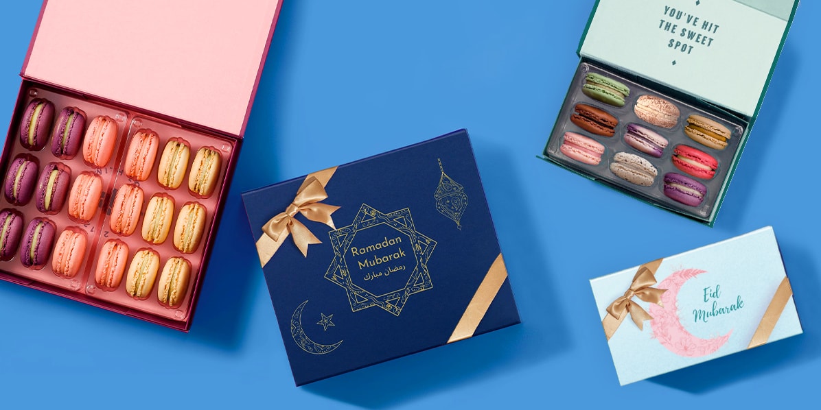 Box of 9 Woops! macarons in assorted flavors, next to a “Eid Mubarak” blue sleeve with a pink moon and a golden ribbon, and Box of 18 Honey Lavender, Rose, and Vanilla French macarons, next to a Ramadan Mubarak blue sleeve with a golden ribbon.