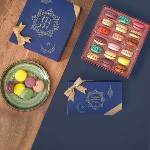 A box full of 18 assorted macarons has a macaron box with a Ramadan sleeve below. To the left is a macaron box with a Ramadan sleeve and a plate full of assorted macarons.