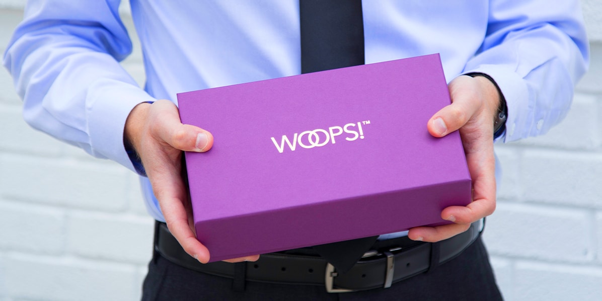 A man in a blue shirt and black pants & tie is holding a purple Woops! box. in his hands.