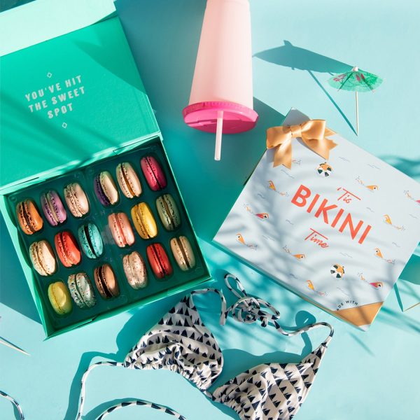 A light blue background features a Woops! box of 18 French macarons with assorted flavors and a macaron box with a “Bikini Time” summer sleeve to its right. A bikini, a pink cup, and some cocktail umbrellas are lying around.