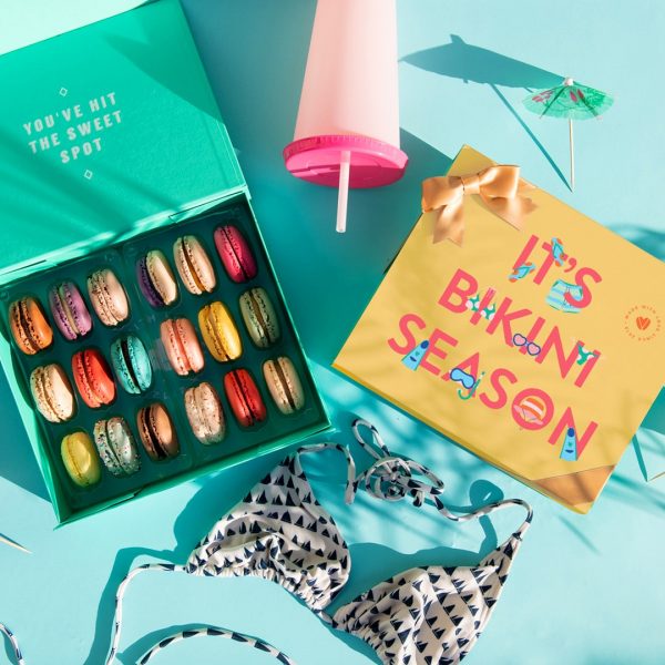 A light blue background features a Woops! box of 18 French macarons with assorted flavors and a macaron box with a “Bikini Time” summer sleeve to its right. A bikini, a pink cup, and some cocktail umbrellas are lying around.