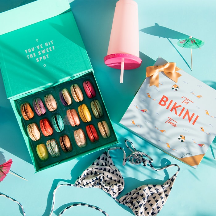 A light blue background features a Woops! Box of 18 French macarons with assorted flavors and a “Bikini Time” summer sleeve to its right. A bikini, a pink cup, and some cocktail umbrellas are lying around.