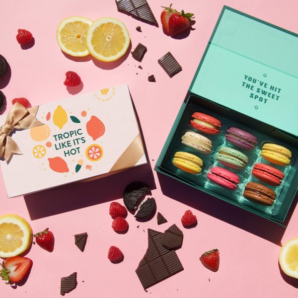 A light pink background features a box of 9 French macarons with assorted flavors and a macaron box with a “Tropic Like It's Hot” summer sleeve to the left. Some fruits and chocolates are lying around.