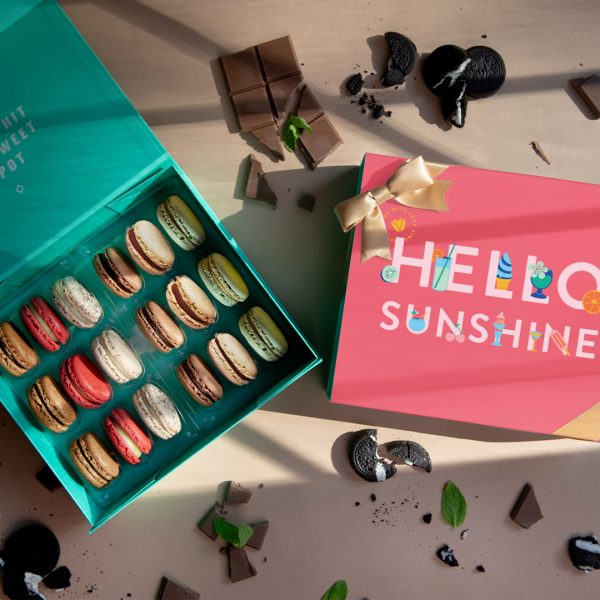 A cream-colored background features a box of 18 French macarons with a Summer sleeve to its right and several chocolates and cookies lying around.