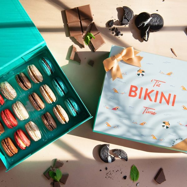 A cream-colored background features a box of 18 French macarons with a “Summer State of Mind” summer sleeve to its right and several chocolates and cookies lying around.