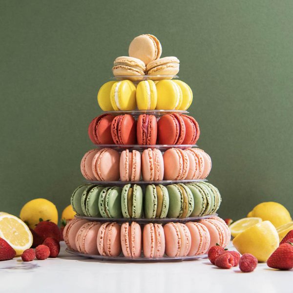 A green background features a large-sized French macaron pyramid with Vanilla, Lemon Meringue, Red Velvet, Strawberry, and Pistachio flavors. Lying around ares slices of lemon, strawberries, and raspberries.