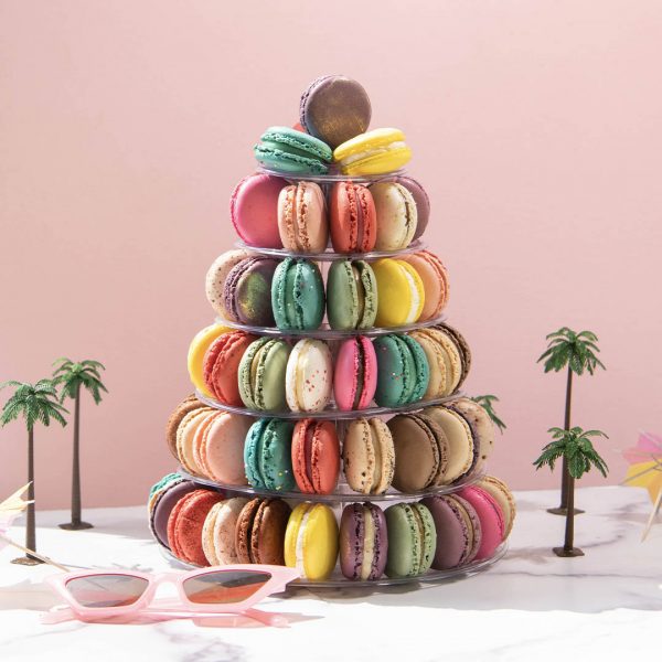 A French macaron pyramid with assorted flavors is surrounded by small palm trees, cocktail umbrellas, and a pair of sunglasses.
