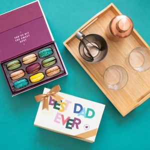 A box full of nine assorted macarons has a macaron box with a Father's Day sleeve below and a wooden table with some cocktail essentials to the right.