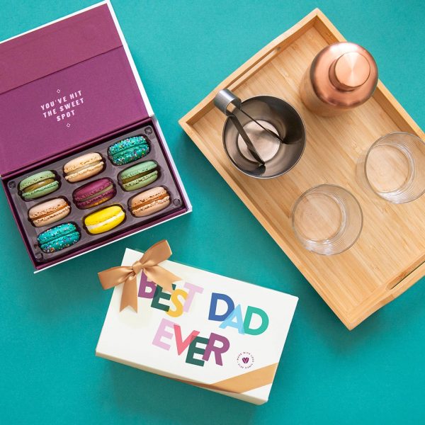 A box full of nine assorted macarons has a macaron box with a Father's Day sleeve below and a wooden table with some cocktail essentials to the right.