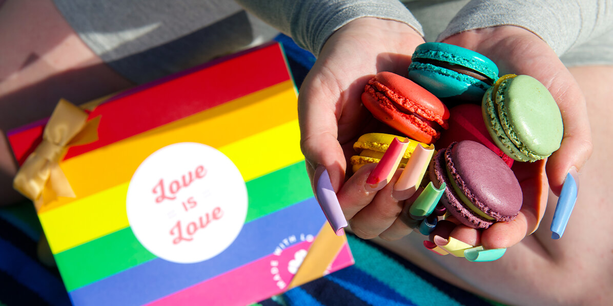 A woman with large, colorful nails is holding a bunch of French macarons in her hands. On her knees is a Woops! Box with a Pride sleeve and a golden ribbon