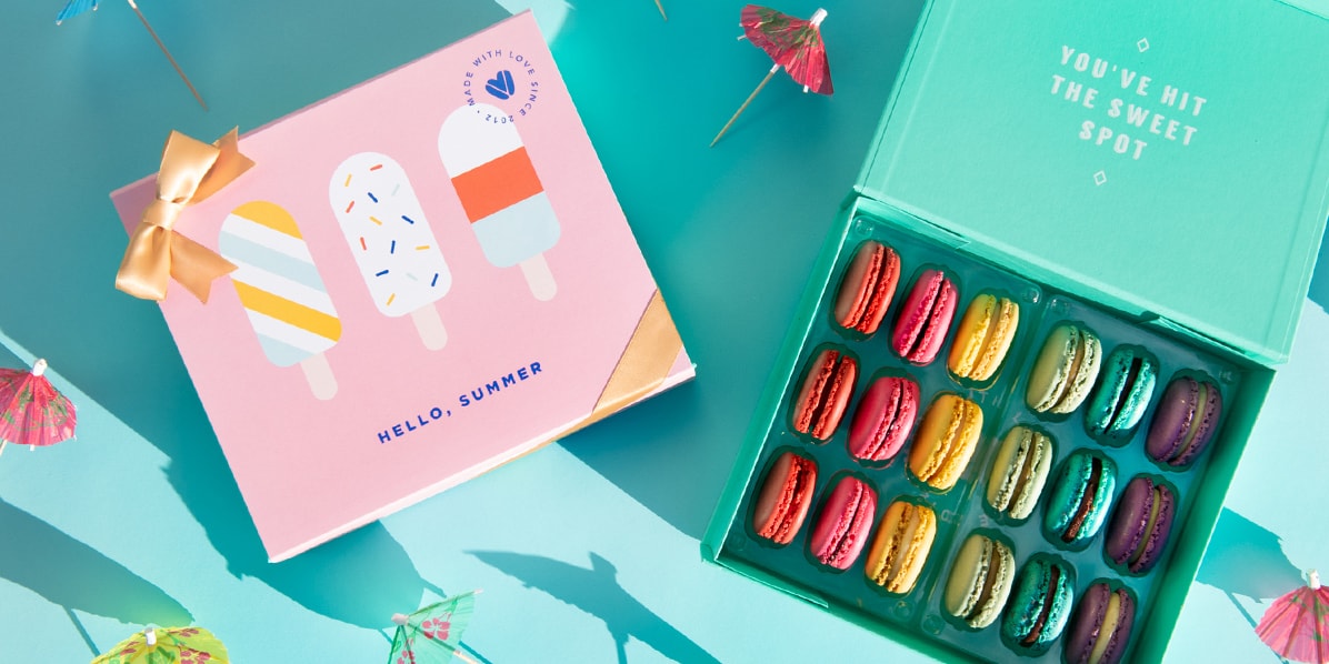 A light blue background features a Woops! Box of 18 French macarons with assorted flavors and a “Hello Summer” summer sleeve to its left. Some cocktail umbrellas are lying around.