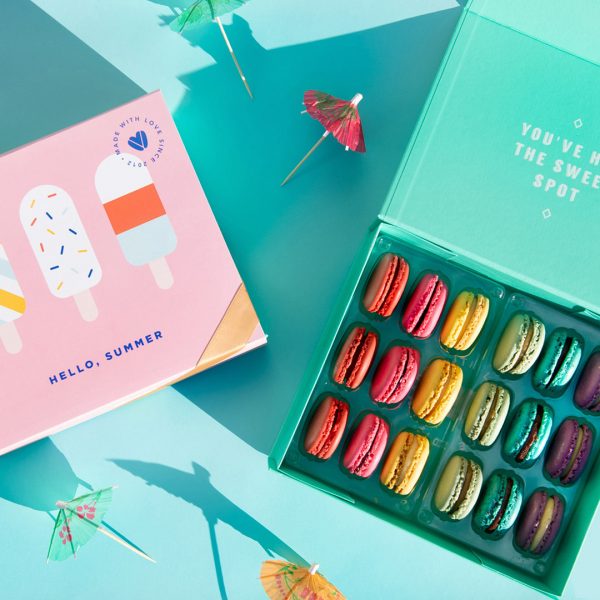 A light blue background features a Woops! box full of 18 assorted French macarons and a macaron box with a “Hello Summer” summer sleeve to its left. Some cocktail umbrellas are lying around.