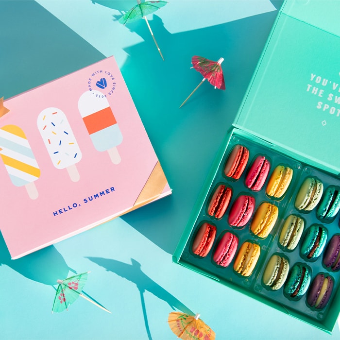 A light blue background features a Woops! Box of 18 French macarons with assorted flavors and a “Hello Summer” sleeve to its left. Some cocktail umbrellas are lying around.