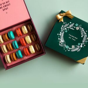 A green background features a Woops! box of 18 French macarons with assorted flavors and a dark green sleeve with a golden ribbon and an ornament figure at the center.