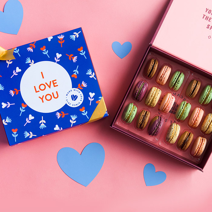 A pink background features a dark blue sleeve with an "I love you" text in the middle and a golden ribbon. To its right is a purple Woops! box of 18 French macarons with assorted flavors and some purple heart figures are lying around.