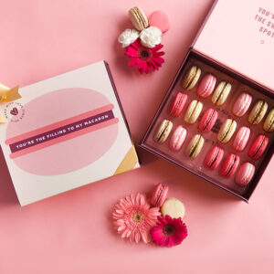 A pink background features a Woops! box that is opened and has 18 French macarons with assorted flavors inside. To its left is a pink sleeve with a French macaron in the middle and a golden ribbon, some flowers with French macarons are lying around.